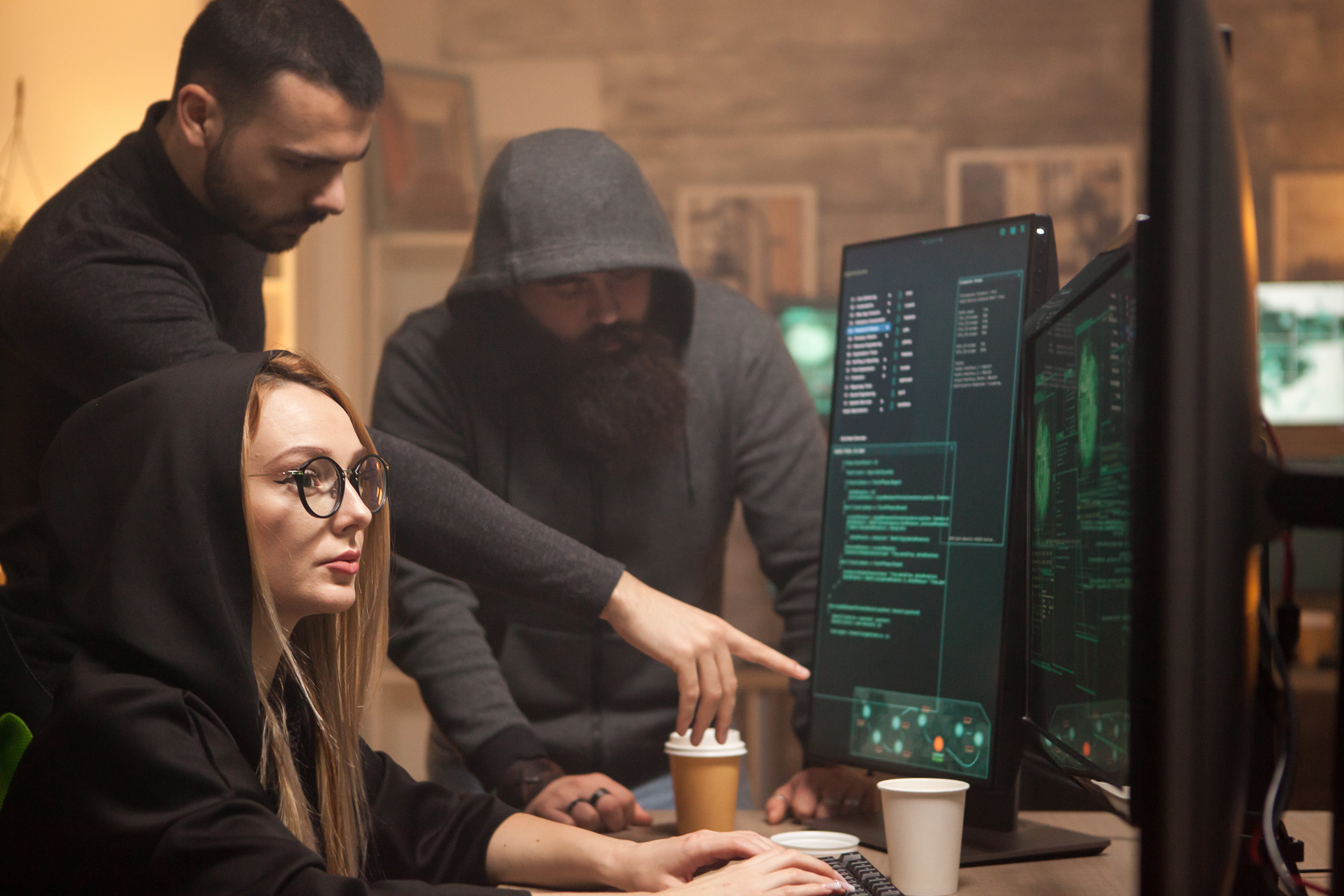 Three people in dark clothing representing cybercriminals looking at a computer screen  